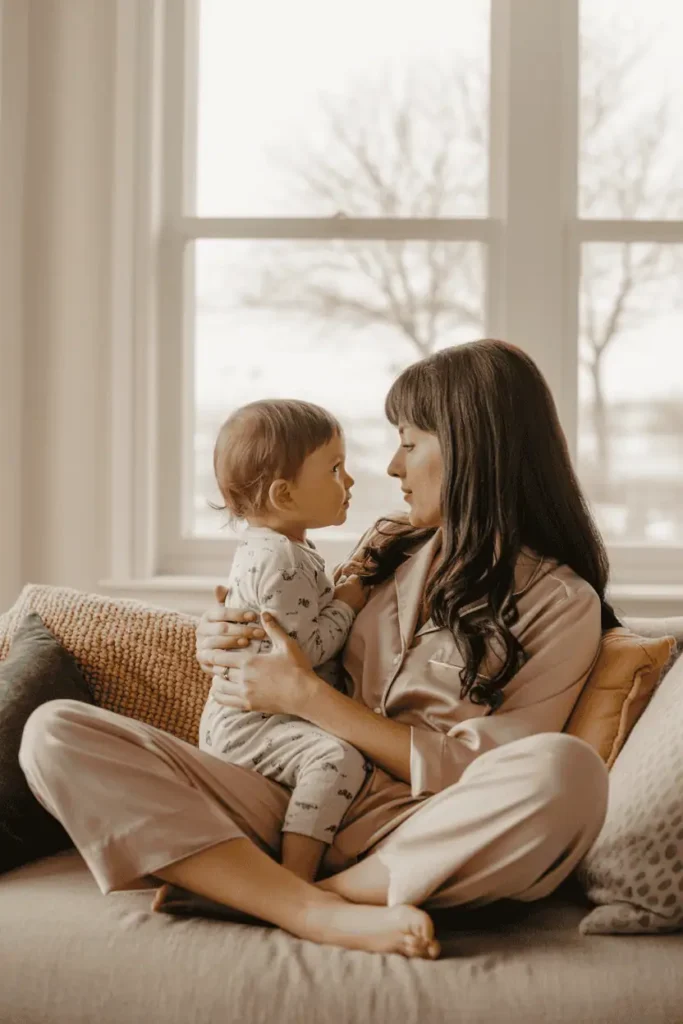 “Candid photo of a mother in pajamas sitting on the couch with a toddler in her lap, soft morning light streaming through a nearby window, warm tones, cozy blankets, natural setting, unfiltered, imperfect and real — relaxed and emotional feel”