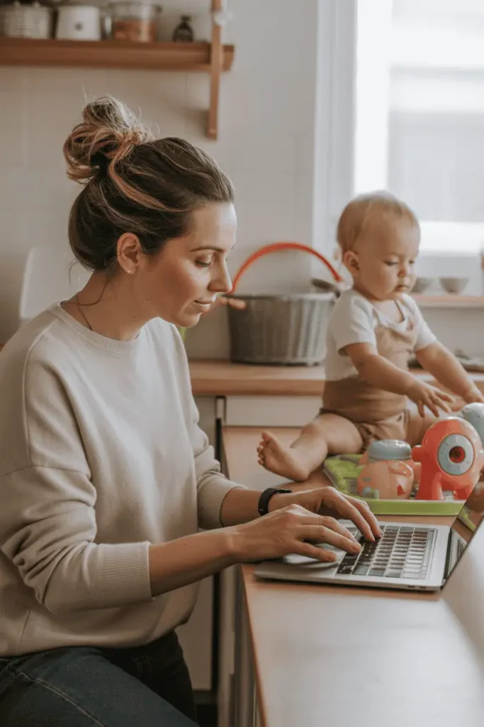 “Mother with messy bun working at laptop in kitchen while toddler plays with toys in the background, natural daylight, neutral tones, organized chaos, cozy productivity mood — real-life work-from-home motherhood”